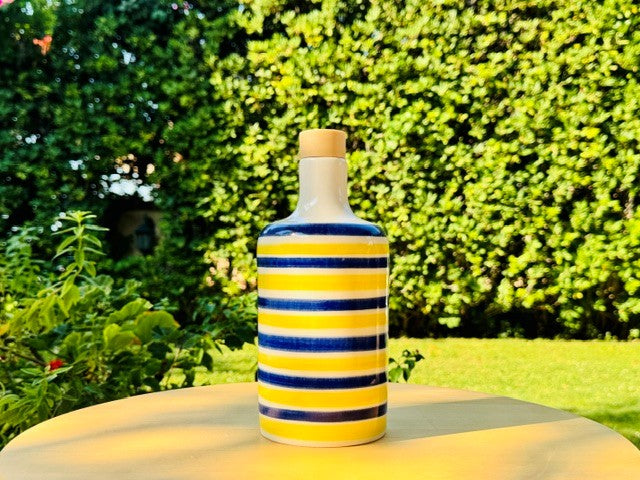 bottiglie in ceramica artigianale decorate a mano con righe blu e gialle vivaci e tappo in legno, perfette come oliere o per l’aceto. Design ispirato alla Costiera Amalfitana, fotografate su un tavolo con foglia di limone, fiori di bouganville e mare sullo sfondo. I colori solari e le forme armoniose donano un tocco d’Italia autentico e raffinato alla cucina, alla tavola o a un angolo esterno mediterraneo.
