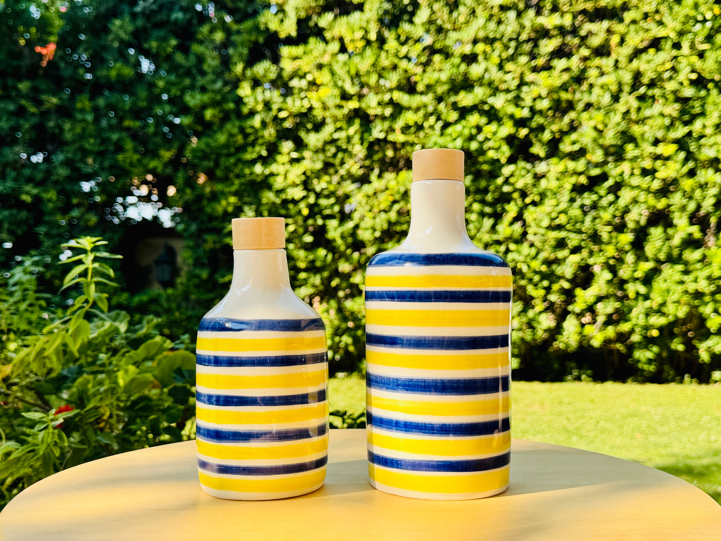 handmade ceramic bottles with bright yellow and blue stripes and wooden caps, perfect as oil or vinegar cruets. Mediterranean design inspired by the Amalfi Coast, placed on a table with a lemon leaf, vibrant bougainvillea, and sea in the background. The vivid colors and elegant shape bring a touch of Italy to your kitchen or dining table—both decorative and functional, ideal for Mediterranean-style homes or outdoor decor.