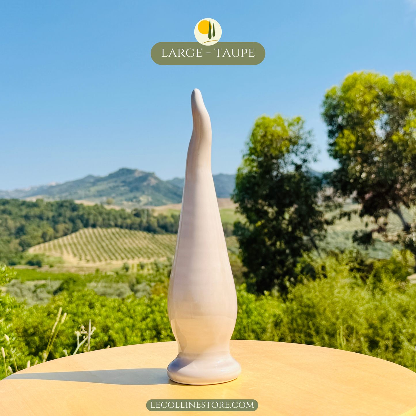 Large taupe ceramic lucky horn with a smooth glossy surface and elongated curved shape. Decorative accent piece for modern home decor, photographed in natural sunlight with a Mediterranean landscape in the background.