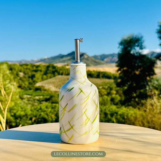 Weiße Keramikflasche mit glänzender Glasur steht auf einem runden, hellen Tisch im Freien. Die Flasche ist mit einem Edelstahl-Ausgießer versehen und mit feinen, handgemalten grünen Pinselstrichen dekoriert, die an Olivenzweige erinnern. Die Oberfläche reflektiert das warme Sonnenlicht. Im Hintergrund sind Olivenbäume, sanfte Hügel und ein klarer blauer Himmel zu sehen.