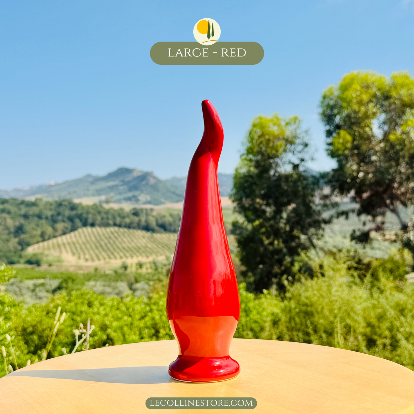 Großes handgefertigtes Keramik chili peperoncino mit glänzend roter Glasur auf hellem Tisch vor mediterraner Landschaft mit Olivenheinen und Hügeln.