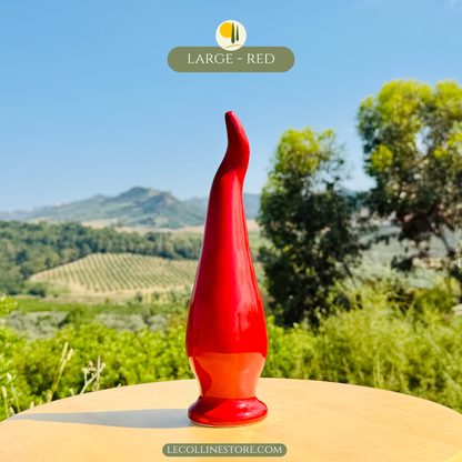Großes handgefertigtes Keramik chili peperoncino mit glänzend roter Glasur auf hellem Tisch vor mediterraner Landschaft mit Olivenheinen und Hügeln.