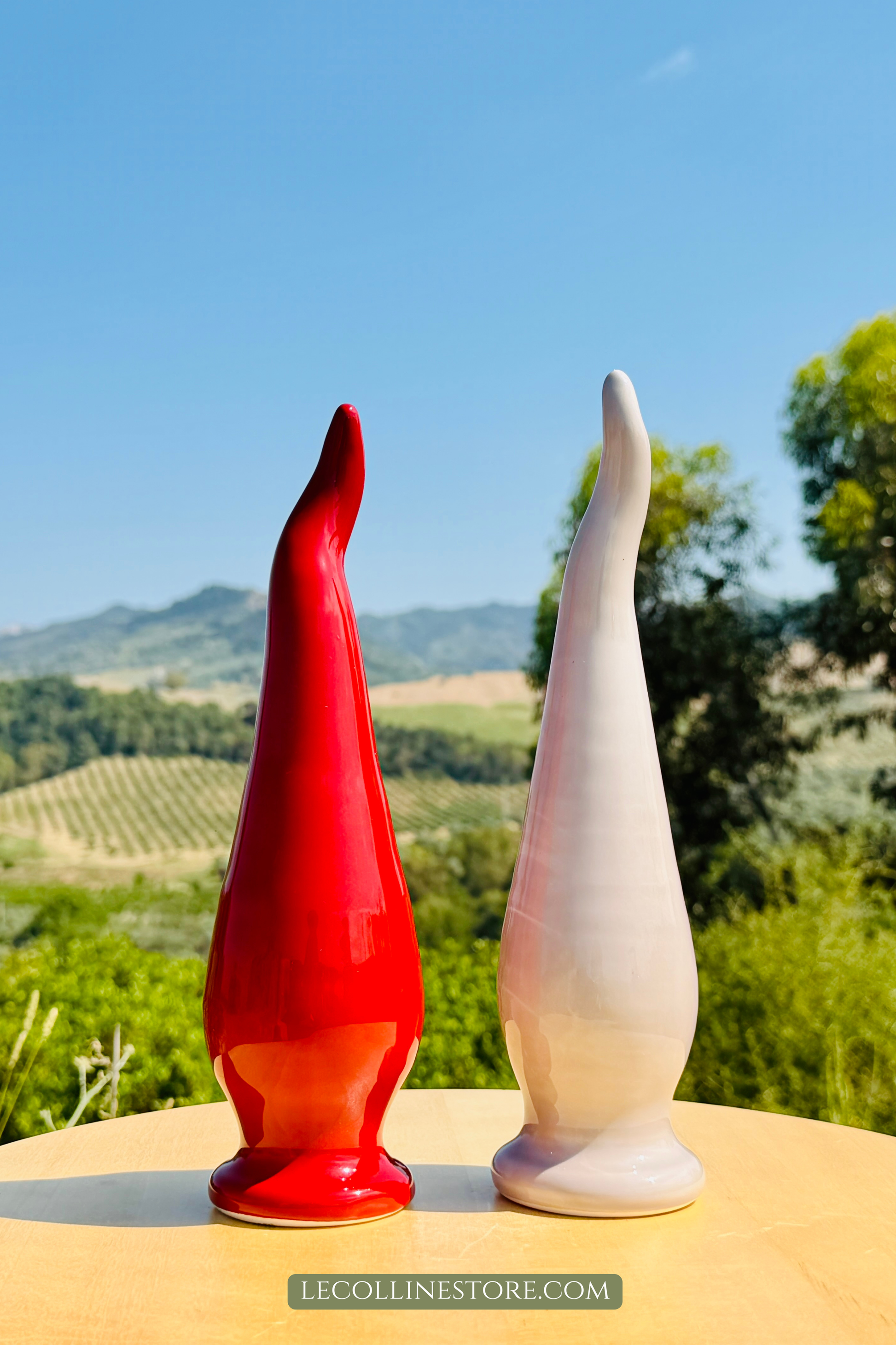 Set of two handcrafted ceramic lucky peperoncino chili pepper horns in glossy red and taupe finishes standing side by side on a light table. Elegant Italian home decor with sculptural forms, displayed against vineyards and green hills.