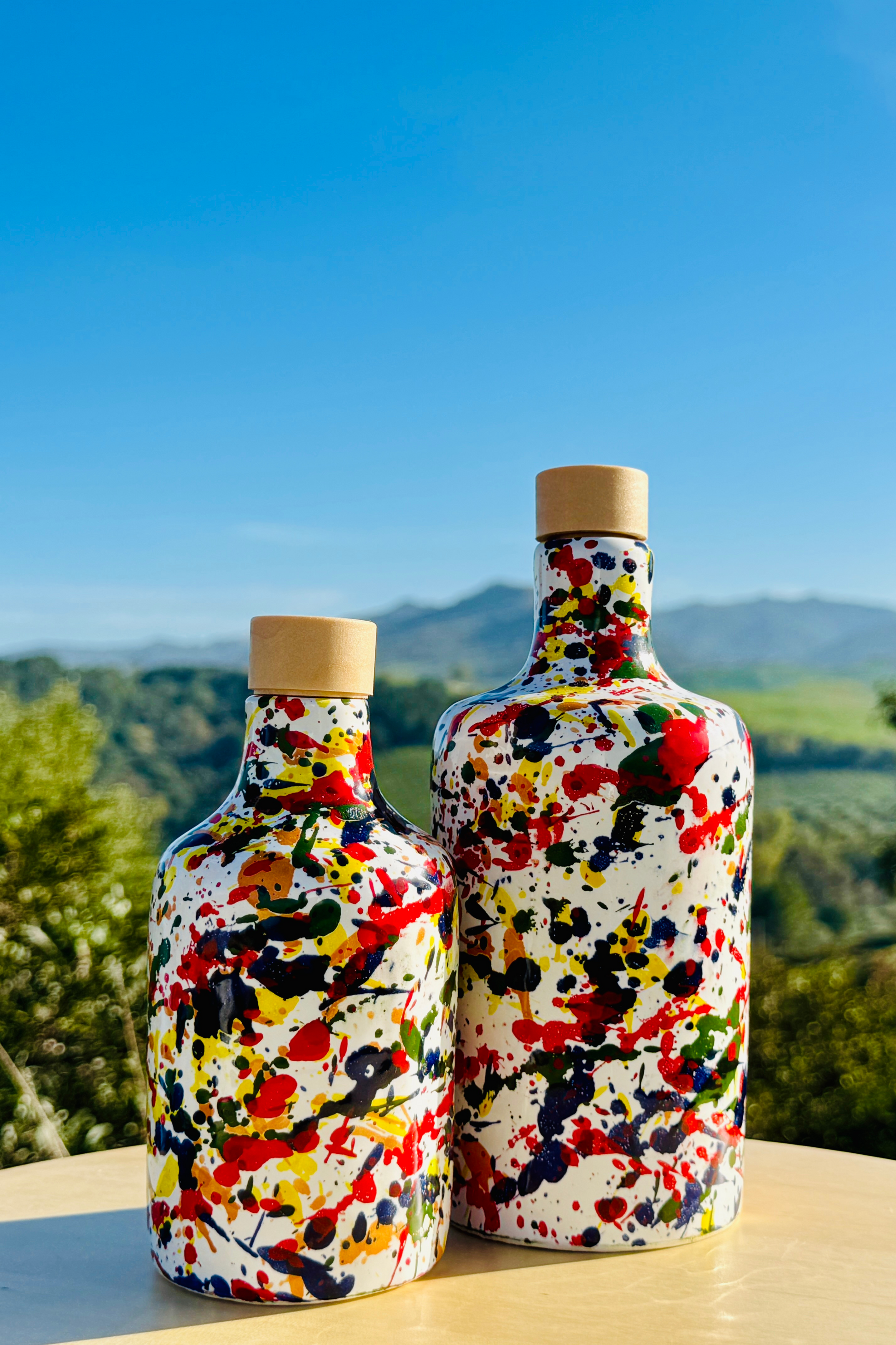 Zwei handbemalte Keramikflaschen im farbenfrohen Splatter-Dekor mit Korken, inszeniert in einer mediterranen Olivenlandschaft in Süditalien