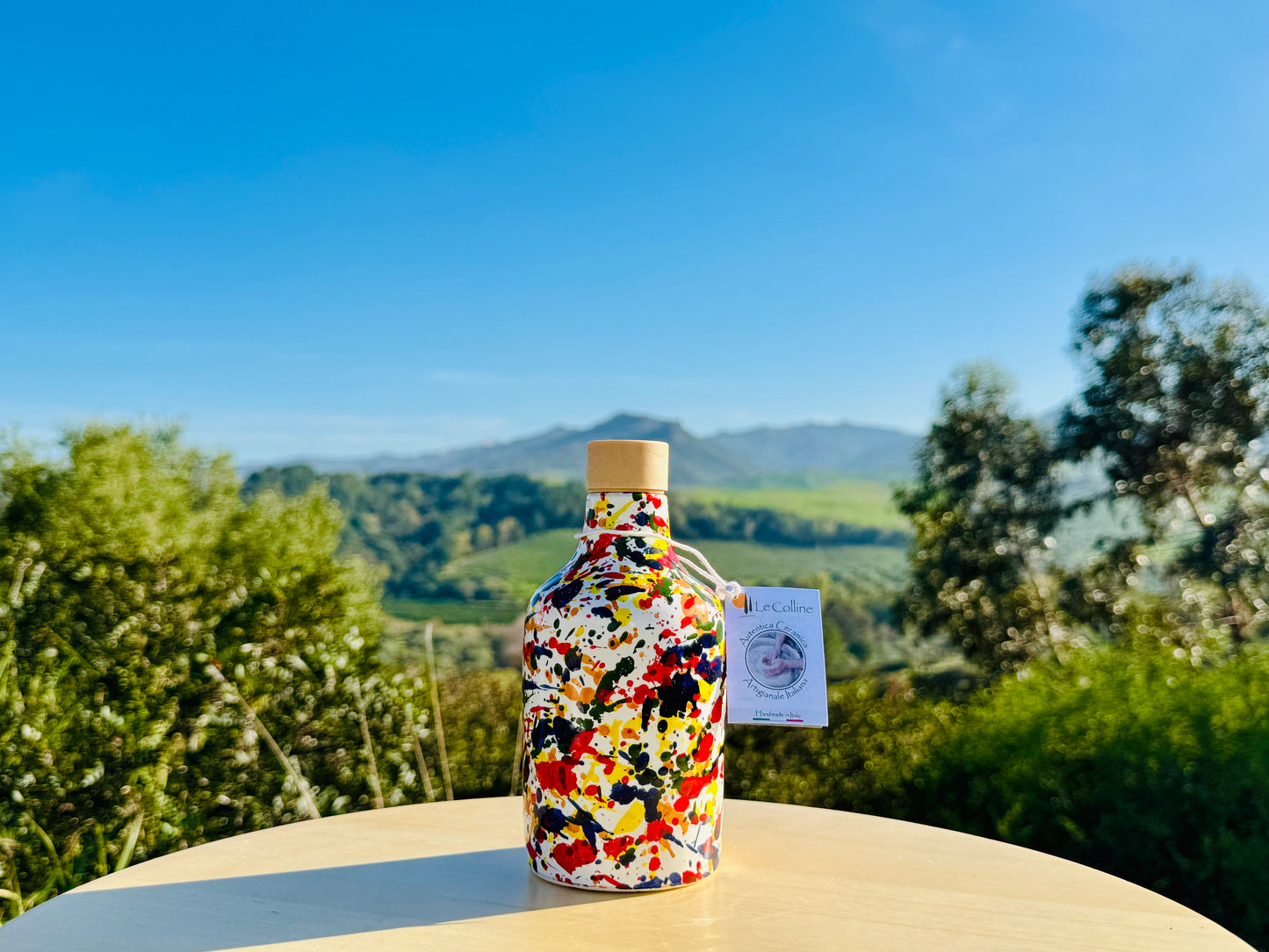 Handbemalte Keramikflaschen 250ml im farbenfrohen Splatter-Dekor mit Ausgießer, inszeniert in einer mediterranen Olivenlandschaft in Süditalien