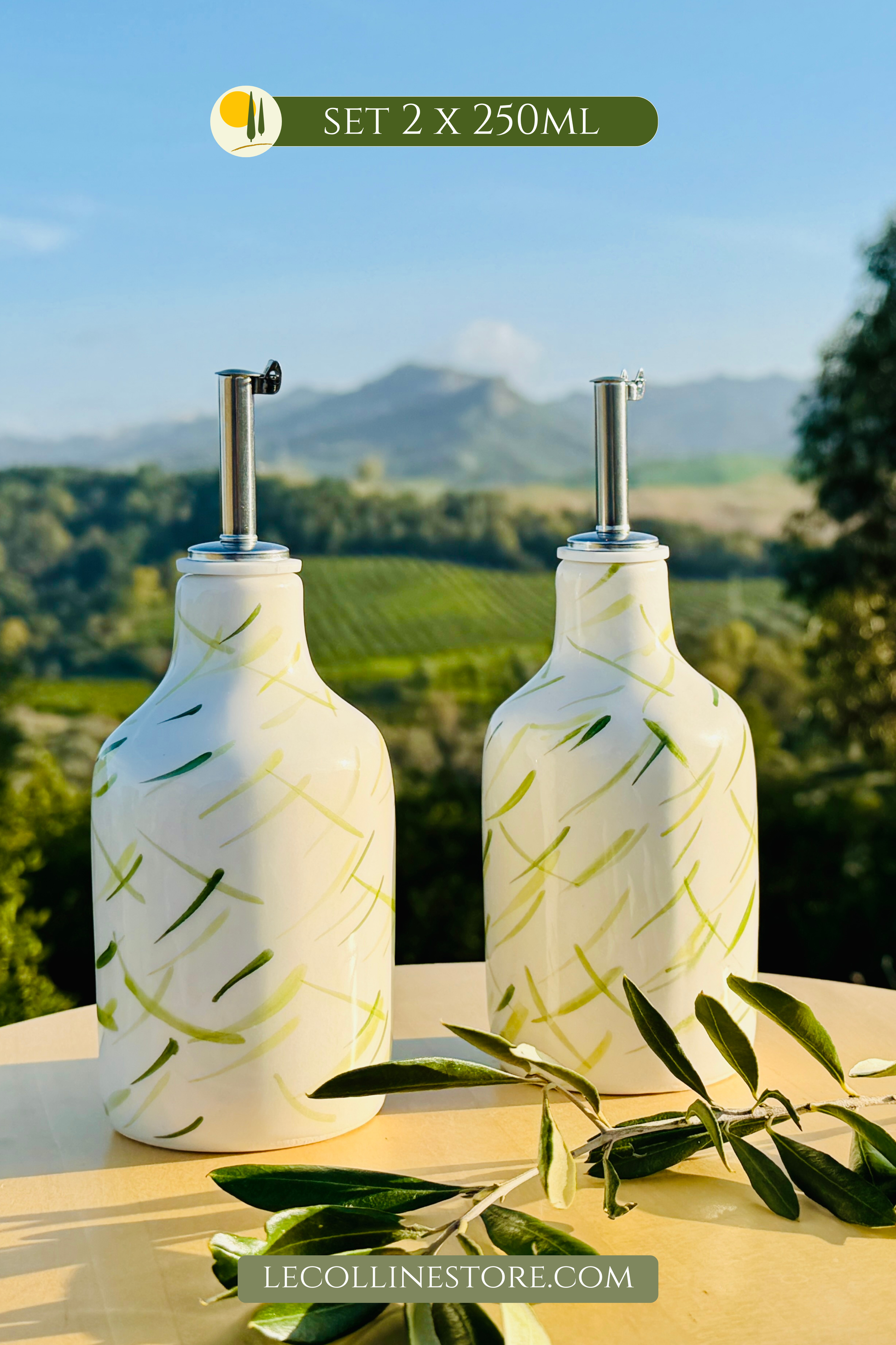 Zwei weiße Keramikflaschen mit glänzender Glasur stehen nebeneinander auf einem runden, hellen Tisch im Freien. Beide Flaschen sind mit einem Edelstahl-Ausgießer versehen und mit feinen, handgemalten grünen Pinselstrichen dekoriert, die an Olivenzweige erinnern. Die Oberfläche reflektiert das warme Sonnenlicht. Vor den Flaschen liegt ein echter Olivenzweig. Im Hintergrund sind Olivenbäume, sanfte Hügel und ein klarer blauer Himmel zu sehen.